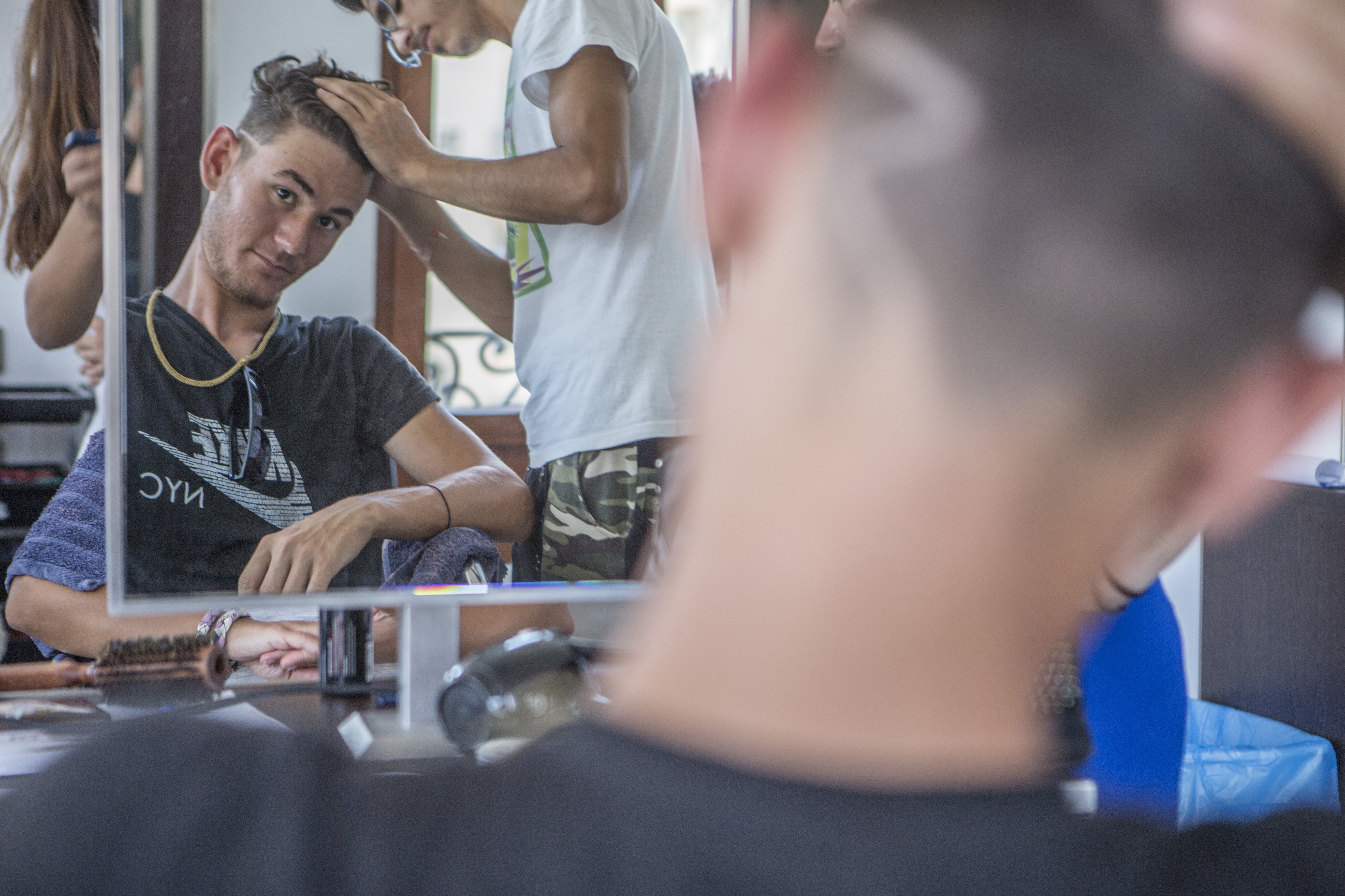 A man sits in a barber chair, looking into a mirror, while having his head shaved.