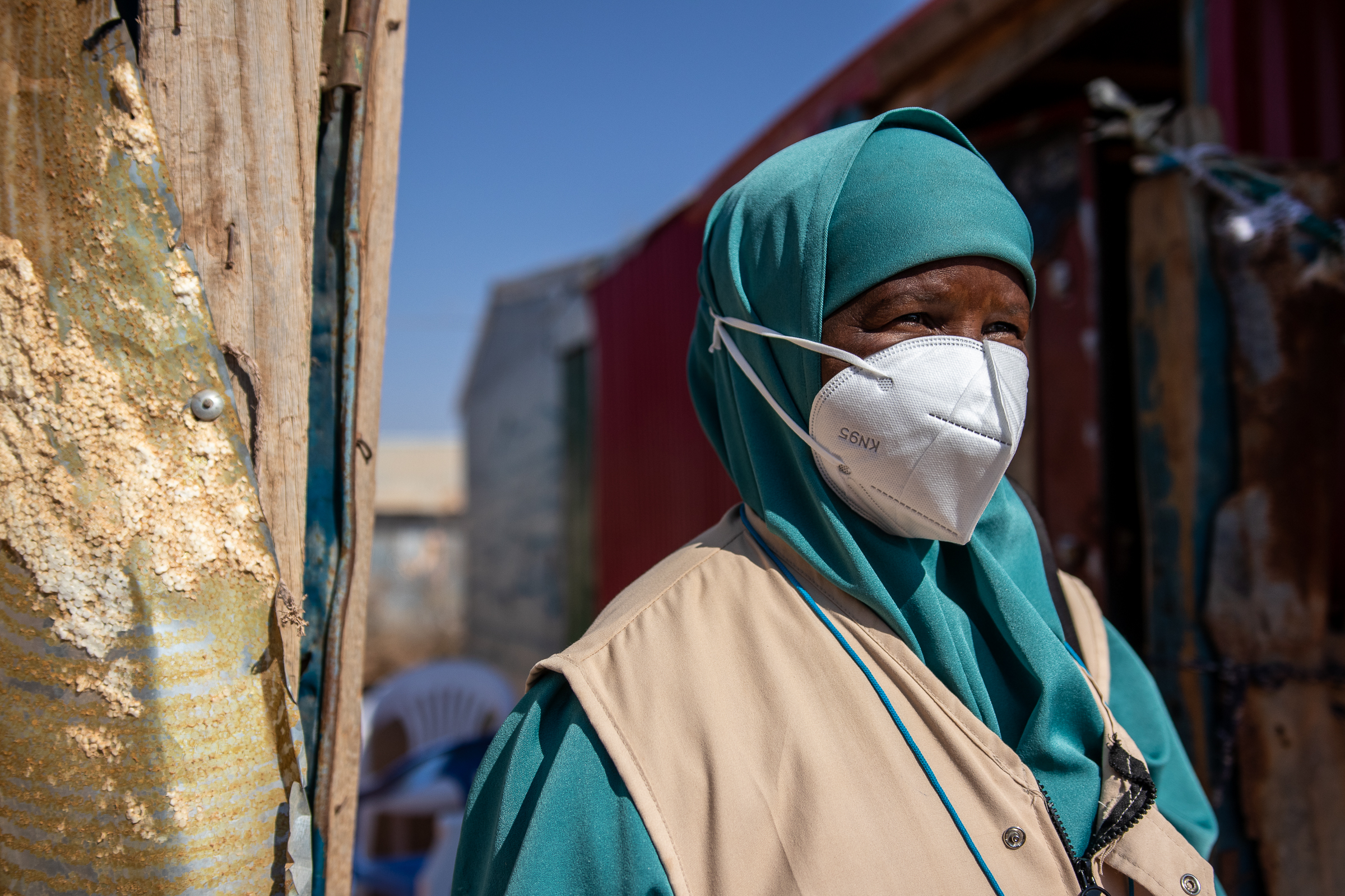 A woman standing outside by a wall wears a face mask and looks at the camera. She is wearing a turquoise coloured uniform and head covering, and a beige vest.