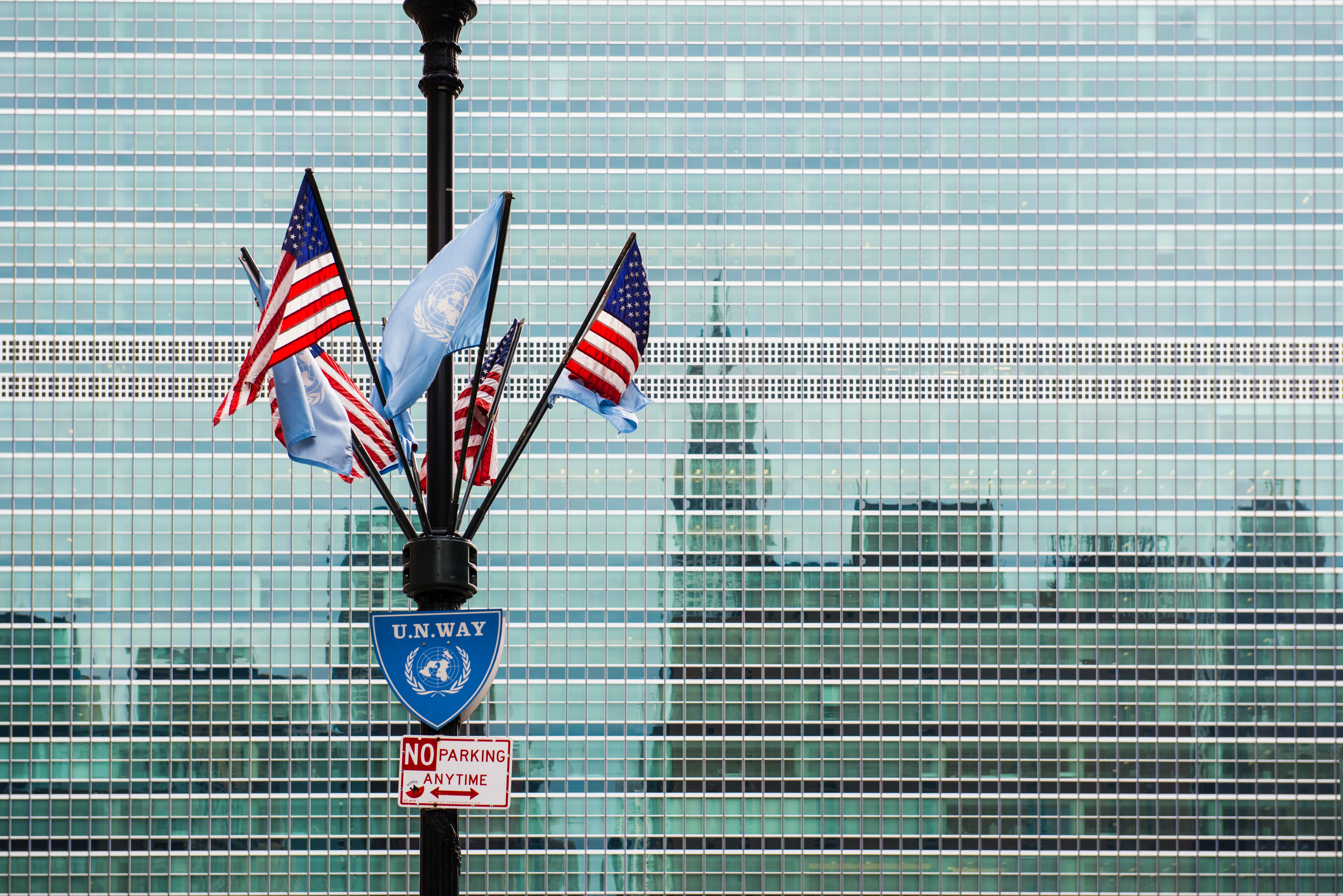 A pole with multiple American and UN flags hangind from it is positioned in front of a building with a reflective facade. The reflection of a city skyline is displayed on the building.