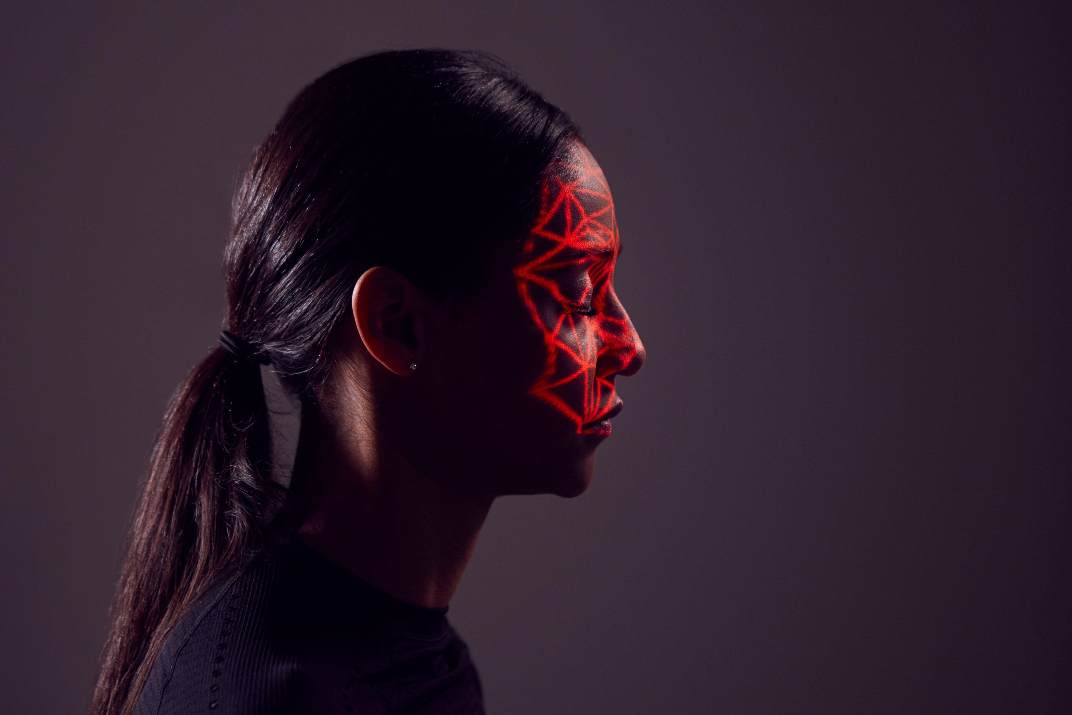 A woman with a black top and hair in a ponytail faces sideways. Her eyes are closed and a red light casts a web-like light on her face.